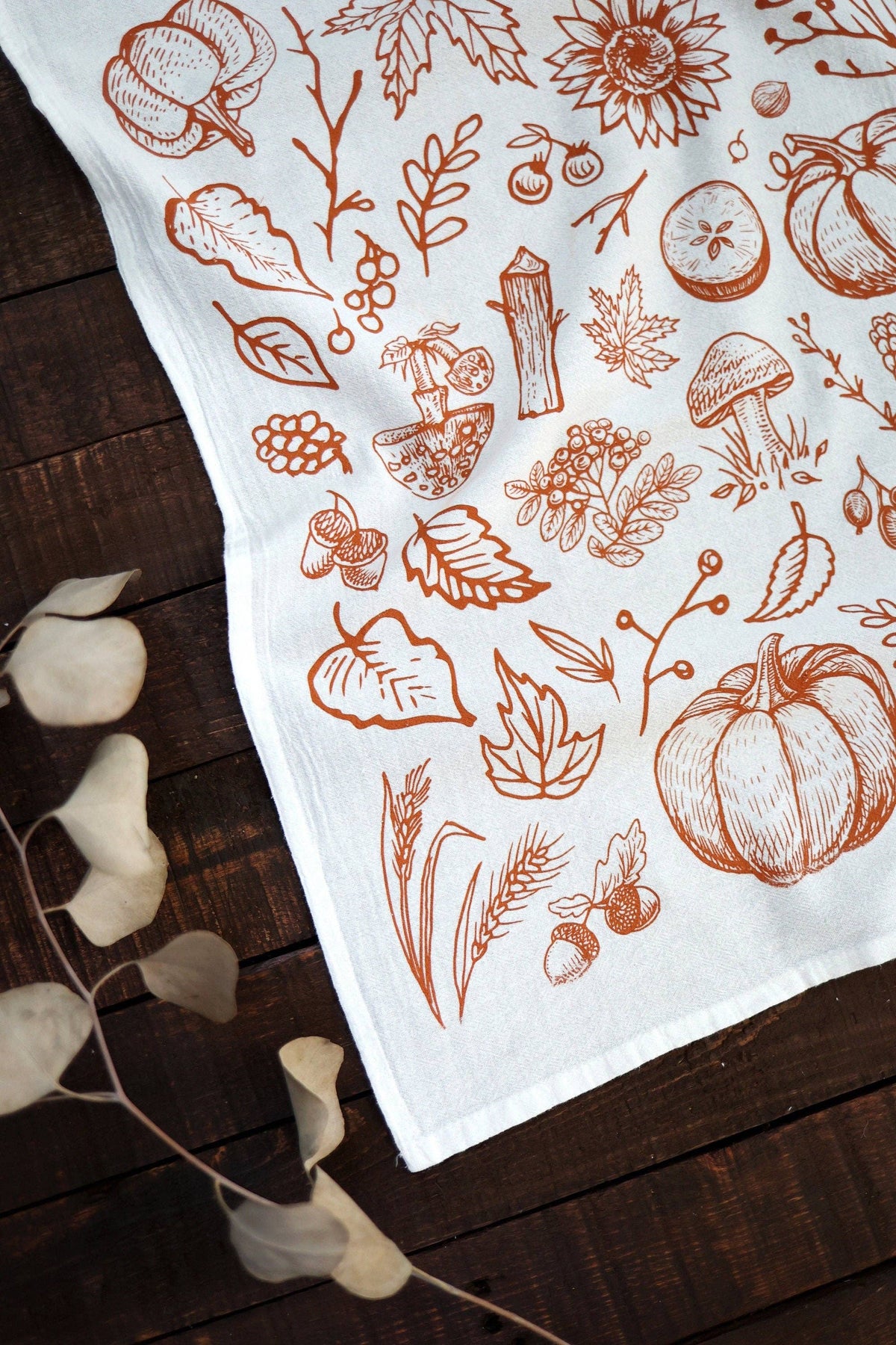 A white fabric tea towel with a brown autumn-themed line art print of pumpkins, leaves, acorns, and mushrooms, styled on a dark wooden surface with dried eucalyptus leaves.