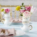 Teacups with floral arrangements on a table setting with pink napkins and gold cutlery.