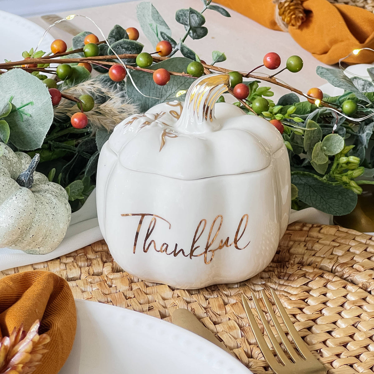 White pumpkin-shaped container with 'Thankful' text on a table setting with greenery and pumpkins.