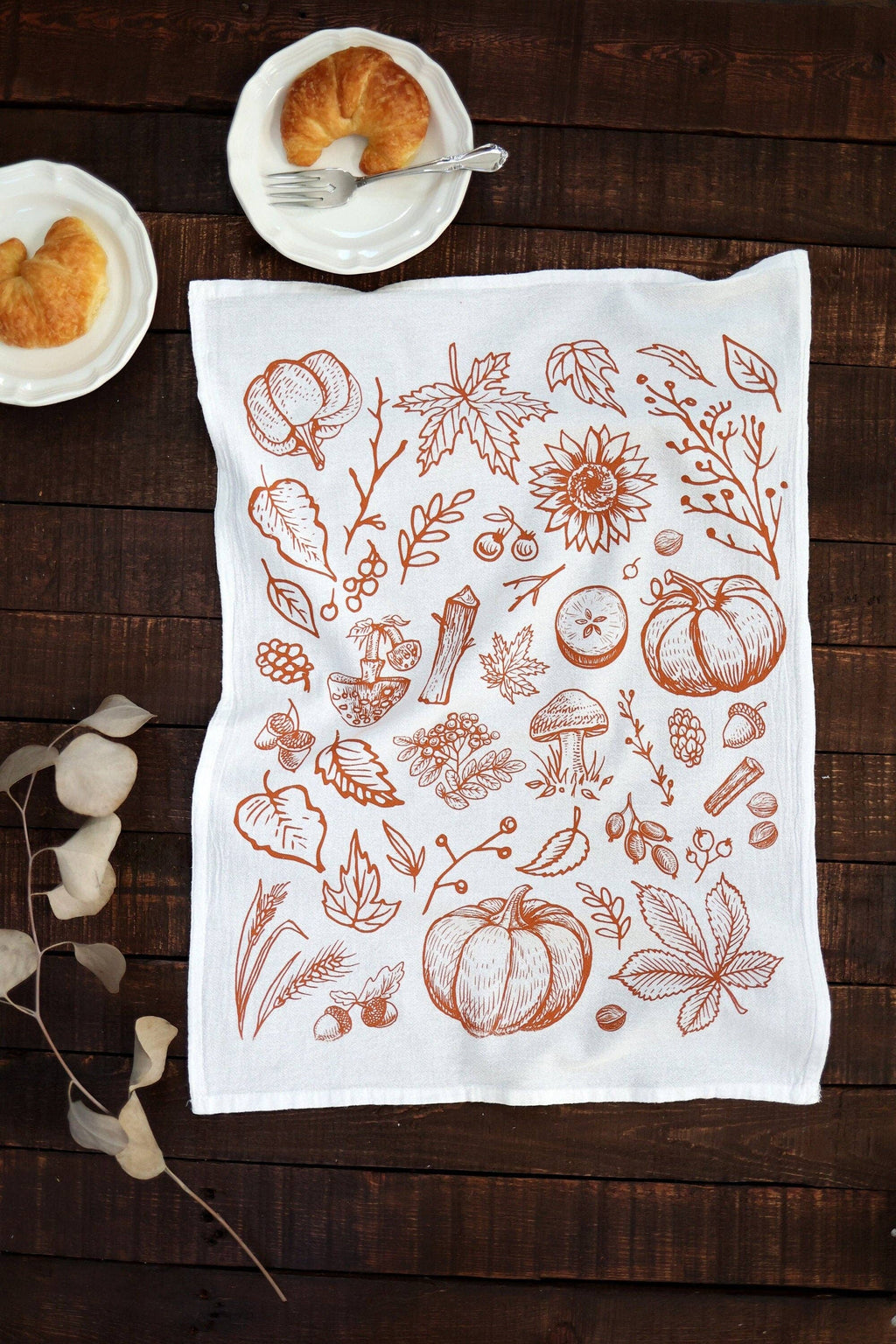 A white kitchen tea towel with a brown autumn-themed line art print of pumpkins, leaves, and mushrooms, laid flat on a dark wooden table with croissants on small white plates.