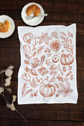 A white kitchen tea towel with a brown autumn-themed line art print of pumpkins, leaves, and mushrooms, laid flat on a dark wooden table with croissants on small white plates.