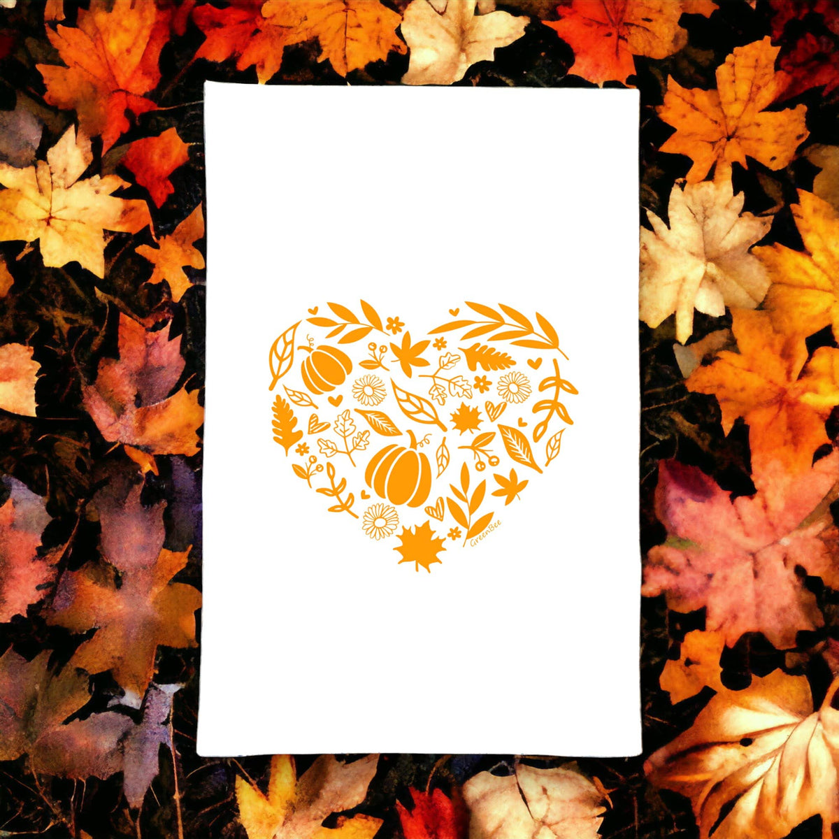A white rectangular tea towel or napkin laid flat, featuring an orange heart-shaped floral and autumn leaf pattern in the center. The towel is photographed from above and is surrounded by a border of colorful fall leaves in orange, yellow, red, and brown.