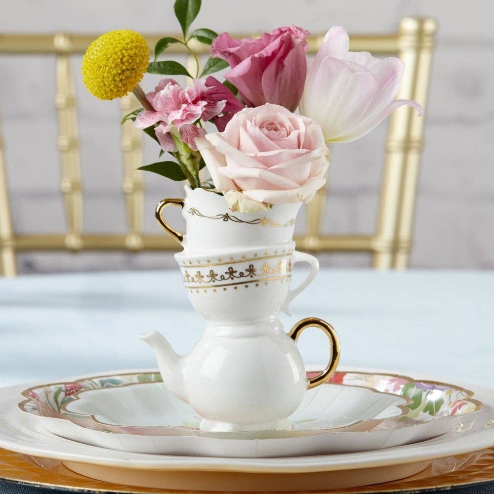 A white porcelain teapot-shaped vase with gold trim and a floral saucer, holding pink and white flowers, set on a table with stacked plates and a gold chair in the background.