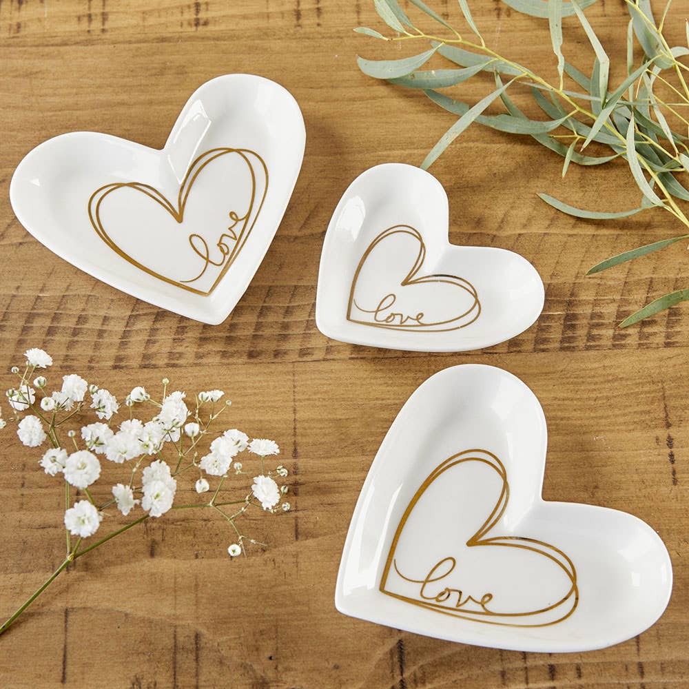Three white ceramic heart-shaped dish holders with gold 'love' script, arranged on a wooden tabletop with white baby's breath and green eucalyptus sprigs.