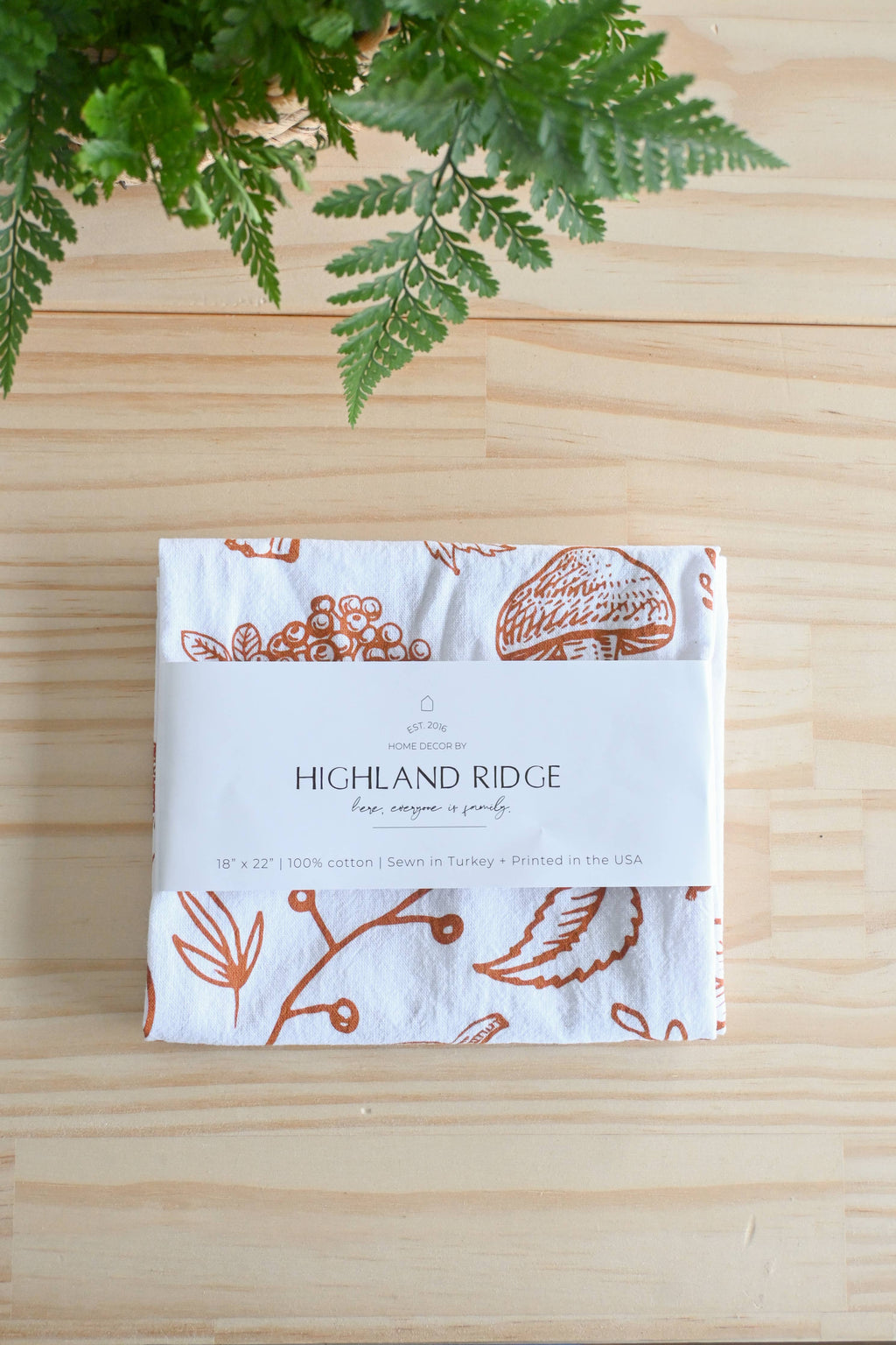 A folded white cotton napkin with rust-orange botanical illustrations and a white paper band reading 'HIGHLAND RIDGE' and 'Highland Ridge Napkins', placed on a light wood surface with green fern leaves at the top.