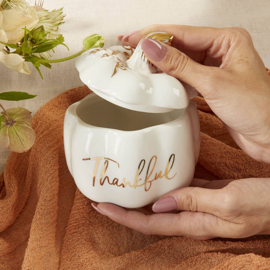 White ceramic mug with 'Thankful' text, held by a hand on a brown fabric background with flowers.
